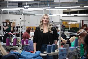 Samantha Gaunt stands on a factory floor. There are sewing machinists and spools of thread working around her.