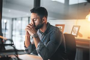 A man sitting at a desk with his elbows on the desk, leaning his head on his forefingers with his eyes closed