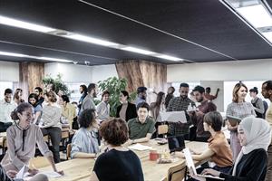 A big office room with a desk containing a lot of people, all too close to each other, some are carrying laptops and paperwork.
