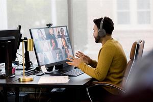 A man wearing headphones sitting at a desk with a computer, taking part in a remote meeting