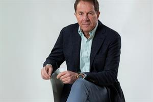 Peter Cheese sits on a chair, leaning forward with one leg crossed over the other and holding a pair of glasses