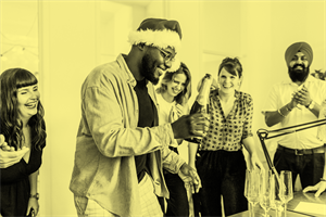 People stood laughing around someone wearing a Santa hat and holding a bottle of Champagne