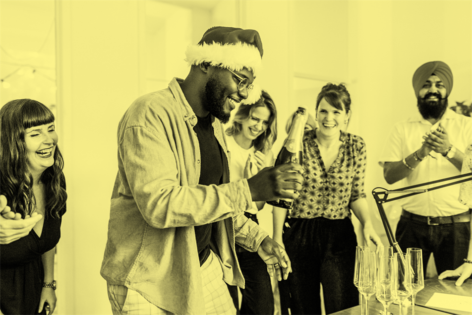 People stood laughing around someone wearing a Santa hat and holding a bottle of Champagne