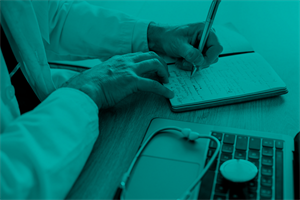 Close up of someone writing in a notepad next to a laptop and a stethoscope