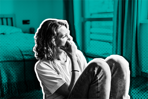 Woman sat next to a bed with knees drawn up and hand on her face