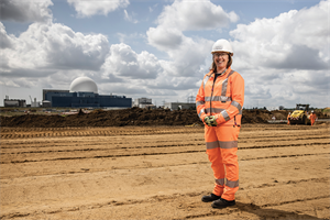 Sarah Hancock wears a white hard had and orange fluorecent PPE suit. She is standing on a construction site with building works in the background